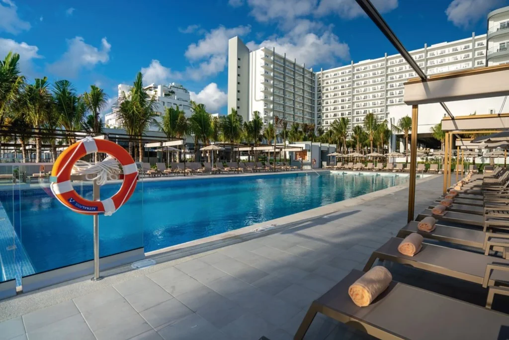 Spacious outdoor swimming pool surrounded by sun loungers, palm trees and modern resort buildings at Riu Palace Kukulkan in Cancun.