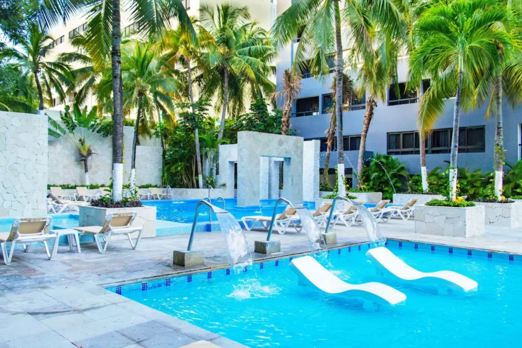 Tropical outdoor swimming pool with waterfall features, submerged loungers and surrounding palm trees at Smart Cancun by Oasis in Cancun.