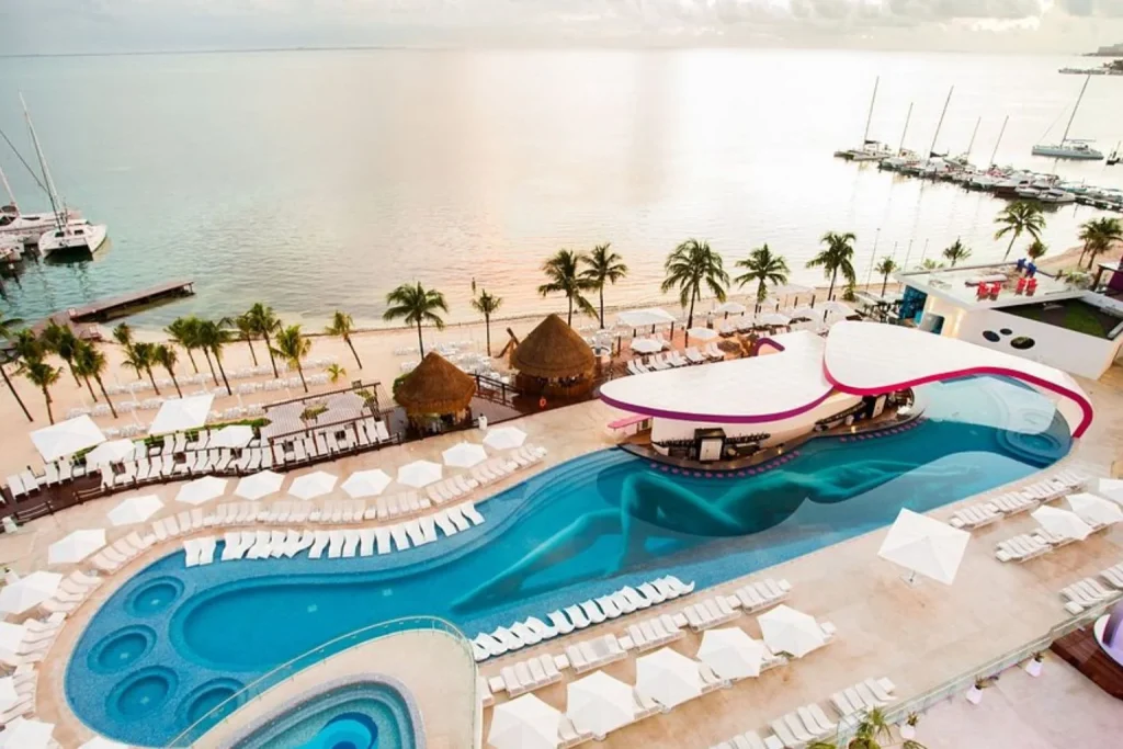 Aerial view of the lagoon-style swimming pool, beachfront sun loungers, private marina, and calm Caribbean waters at Temptation Cancun Resort in Cancún, Mexico.